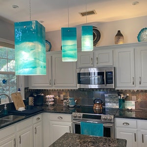 K&ouml;nnte beinhalten: Drei tealblaue Glaspendelleuchten h&auml;ngen von der Decke &uuml;ber einer K&uuml;chentheke. Die Leuchten sind rechteckig und haben einen wei&szlig;en Hintergrund mit einem blauen und wei&szlig;en Marmormuster. Die Leuchten sind eingeschaltet und beleuchten die K&uuml;che.