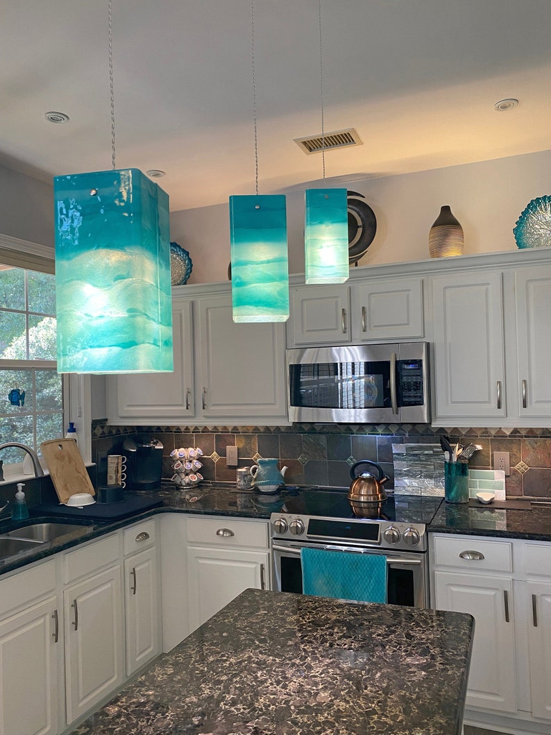 K&ouml;nnte beinhalten: Drei tealblaue Glaspendelleuchten h&auml;ngen von der Decke &uuml;ber einer K&uuml;chentheke. Die Leuchten sind rechteckig und haben einen wei&szlig;en Hintergrund mit einem blauen und wei&szlig;en Marmormuster. Die Leuchten sind eingeschaltet und beleuchten die K&uuml;che.