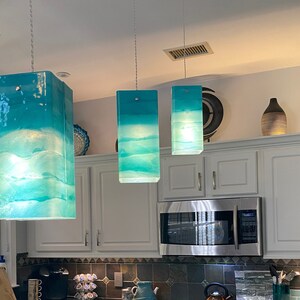 K&ouml;nnte beinhalten: Drei t&uuml;rkisfarbene Glaspendelleuchten mit einem wei&szlig;en und blauen Ombre-Effekt h&auml;ngen von der Decke in einer K&uuml;che. Die Leuchten sind an silbernen Ketten aufgeh&auml;ngt.