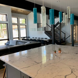 May include: A modern kitchen with a large island featuring a white quartz countertop with grey veining. The island has a grey cabinet with a silver handle.  A set of six teal blue glass pendant lights hang over the island.