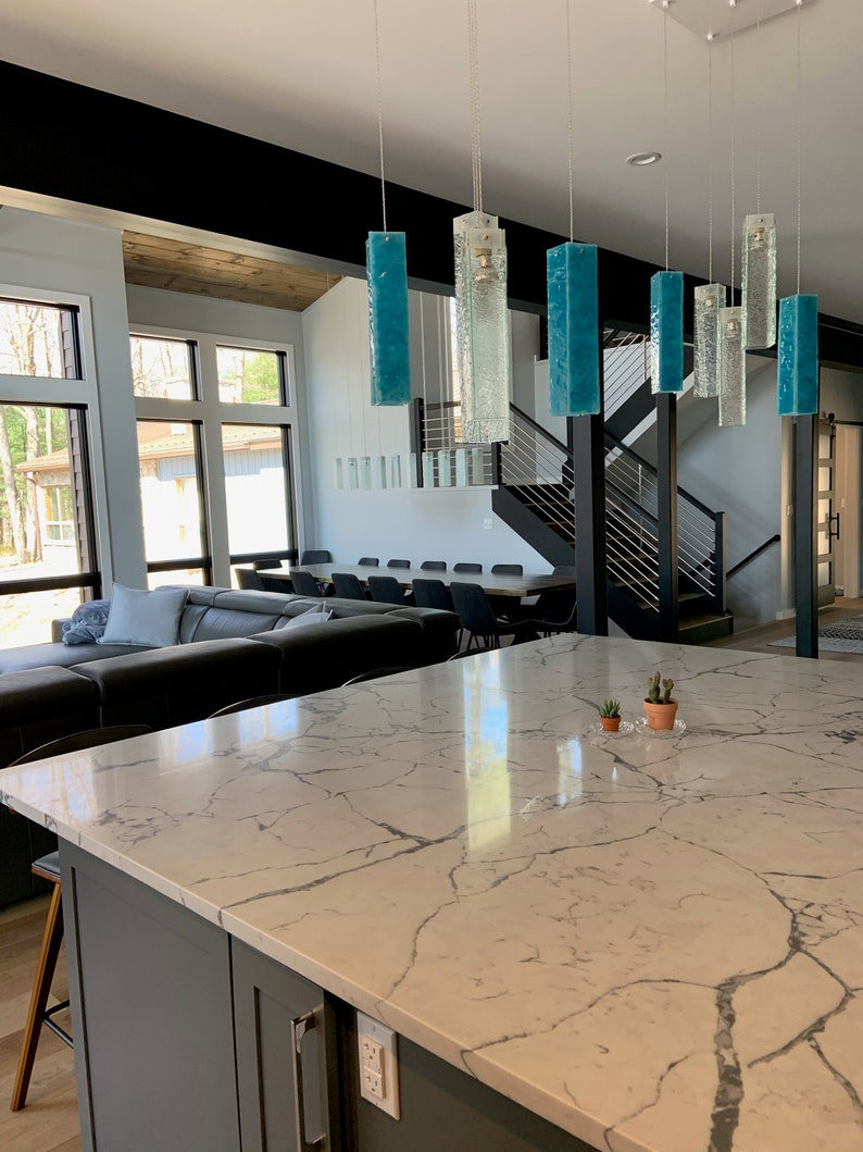 May include: A modern kitchen with a large island featuring a white quartz countertop with grey veining. The island has a grey cabinet with a silver handle.  A set of six teal blue glass pendant lights hang over the island.