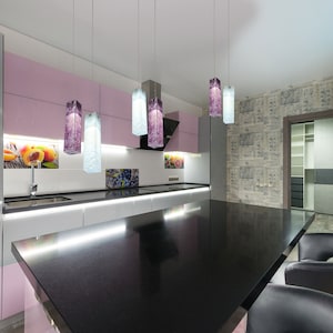 May include: A modern kitchen with a black granite countertop island and bar stools. The kitchen features white cabinets, pink cabinets, and a grey cabinet. The island has a built-in sink and a stainless steel faucet. The kitchen has a modern light fixture with purple and clear glass shades.
