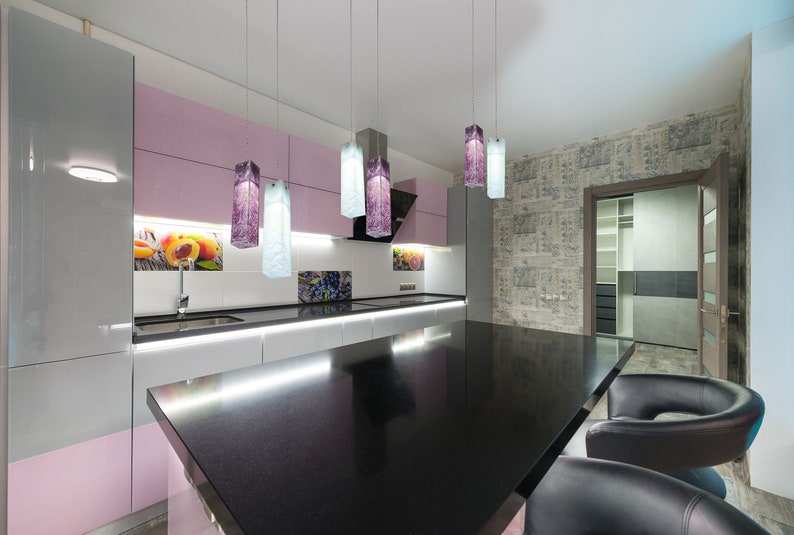 May include: A modern kitchen with a black granite countertop island and bar stools. The kitchen features white cabinets, pink cabinets, and a grey cabinet. The island has a built-in sink and a stainless steel faucet. The kitchen has a modern light fixture with purple and clear glass shades.