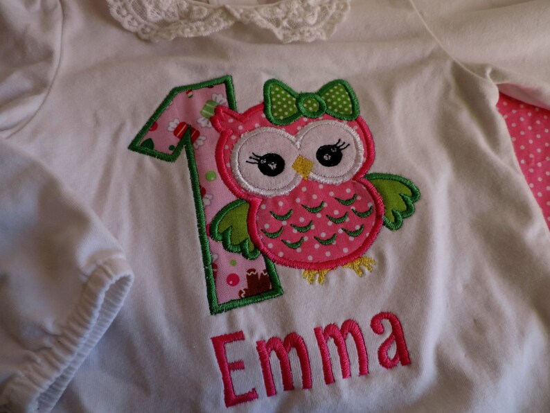 Puede incluir: Camiseta blanca de manga larga con una aplicaci&oacute;n de b&uacute;ho rosa y verde y el n&uacute;mero 1. El nombre "Emma" est&aacute; bordado en rosa debajo del b&uacute;ho.