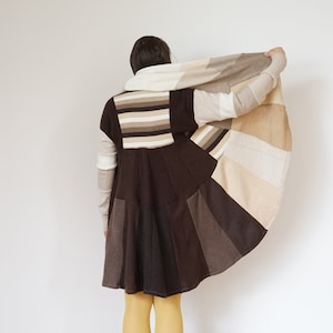 Puede incluir: Un vestido patchwork en tonos marrón, beige y crema, con falda acampanada y mangas largas. El vestido se combina con botas de ante marrones y medias amarillas. La persona sostiene una bufanda a juego.