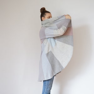 Puede incluir: Un cárdigan gris, rosa y azul con un patrón geométrico. El cárdigan está abierto y drapeado sobre los hombros de la persona.