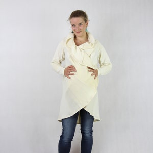 May include: A woman wearing a long, cream-coloured, wrap-style sweater with a cowl neck and long sleeves. She is wearing blue jeans and brown sandals.