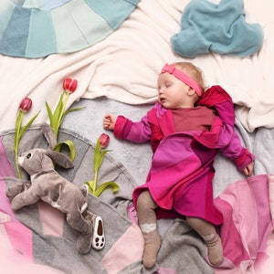 May include: A sleeping baby girl wearing a pink hooded jacket and a pink headband lies on a white and gray blanket with a gray bunny toy and pink flowers.