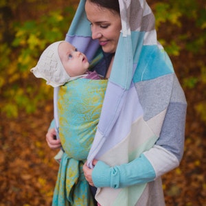 May include: A woman wearing a multi-coloured patchwork hooded cardigan holds a baby wrapped in a turquoise and gold patterned baby wrap. The baby is wearing a white crocheted hat.