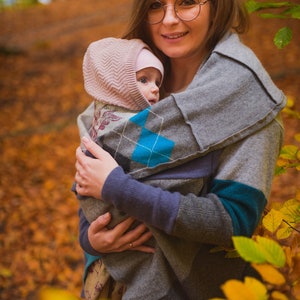 May include: A woman wearing glasses holds a baby wrapped in a grey and blue patterned baby wrap. The baby is wearing a pink hat. The background is a blurry image of autumn foliage.