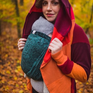 Può includere: Una donna indossa un cardigan con cappuccio multicolore e porta un bambino avvolto in uno sling di colore turchese con motivo. La donna è in piedi in un bosco con foglie autunnali.