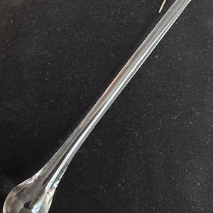 May include: A clear glass teardrop-shaped ornament with a thin, elongated stem and a small, curved gold-colored wire hook. The ornament is transparent, reflecting light, and rests against a dark background.