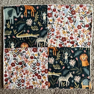 May include: A square baby blanket with a patchwork design. The blanket features four sections with different patterns. Two sections have a dark green background with animal and floral designs. The other two sections have a white background with floral designs.