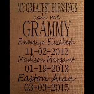 May include: A burlap-textured sign with the words "MY GREATEST BLESSINGS call me GRAMMY" in dark brown lettering. Below are names and dates: Emmalyn Elizabeth (11-02-2012), Madison Margaret (01-19-2013), and Easton Alan (03-03-2015).
