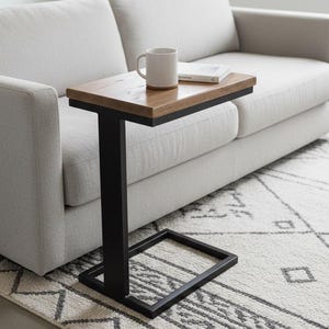 May include: A contemporary C-shaped side table with a wooden top and black metal frame. A white mug and a book rest on the table, next to a light grey sofa. The table is 61 cm long.