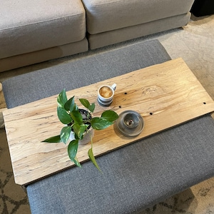 May include: A wooden tray with a light wood grain pattern, featuring a potted plant, a cup of coffee, and a small candle holder. The tray is placed on top of a grey ottoman.