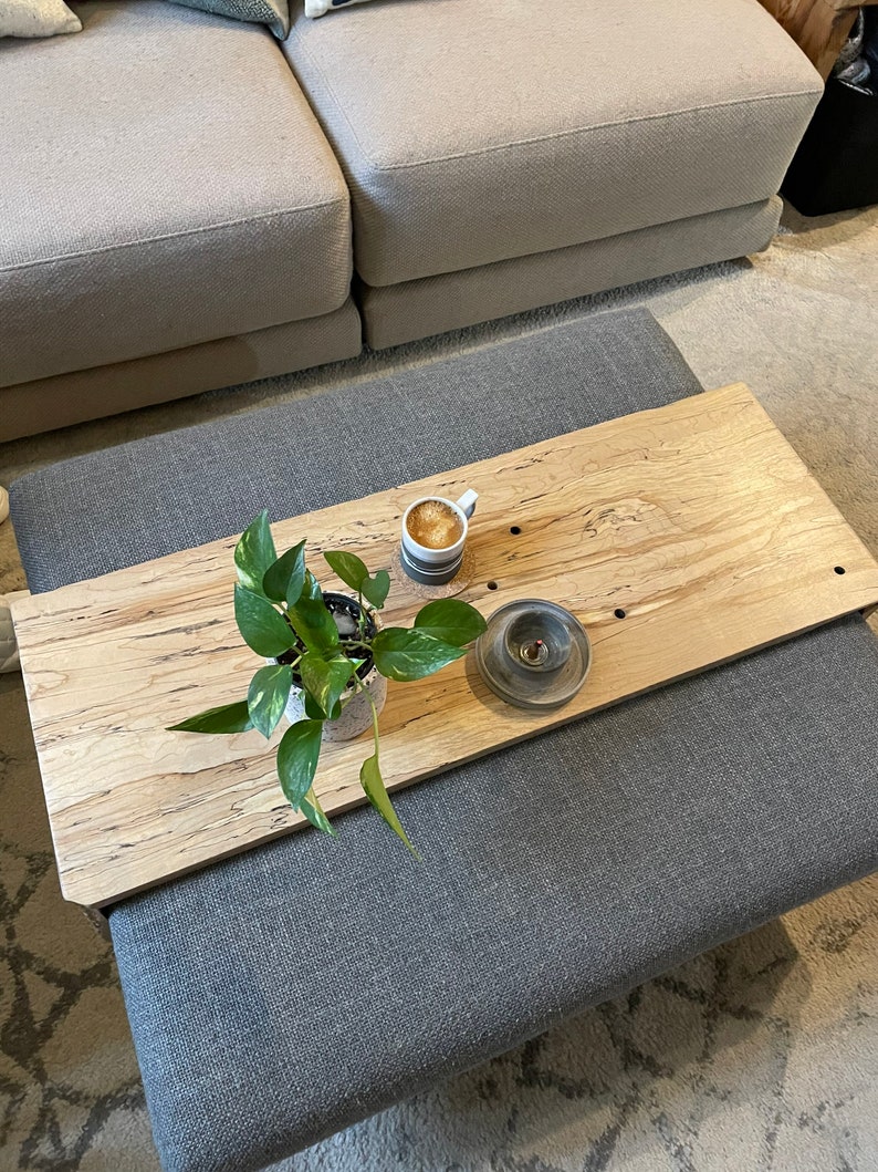 May include: A wooden tray with a light wood grain pattern, featuring a potted plant, a cup of coffee, and a small candle holder. The tray is placed on top of a grey ottoman.