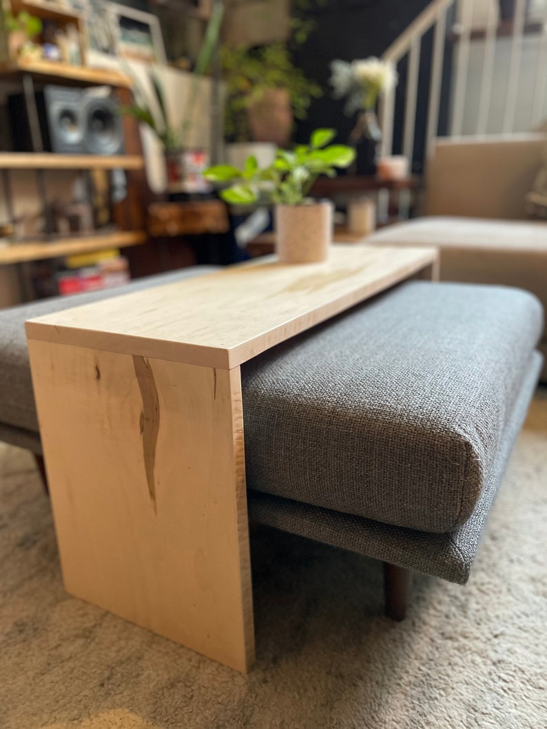 May include: A light wood side table with a gray upholstered ottoman attached. The table has a simple, modern design and is perfect for a living room or bedroom.