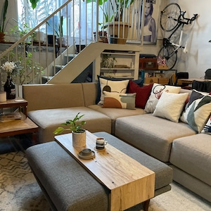 May include: A grey upholstered ottoman with a wooden top that extends out to create a tray. The tray is made of light wood with a natural grain pattern. A small plant and a coffee cup are on the tray. The ottoman is in front of a grey sectional sofa with cushions.