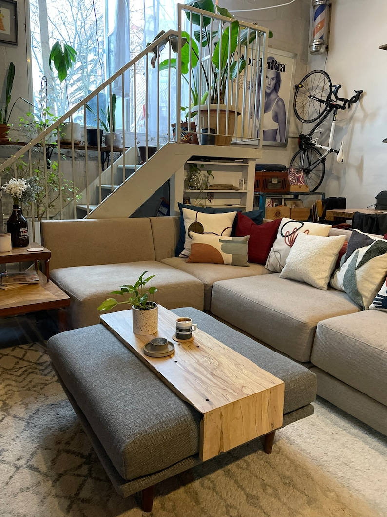 May include: A grey upholstered ottoman with a wooden top that extends out to create a tray. The tray is made of light wood with a natural grain pattern. A small plant and a coffee cup are on the tray. The ottoman is in front of a grey sectional sofa with cushions.