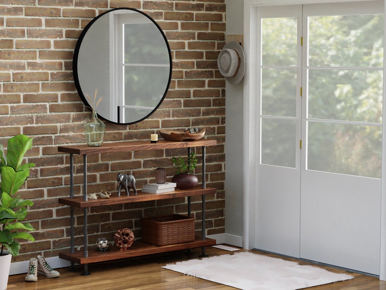 May include: A three-tiered wooden shelf unit with black metal pipe supports. The shelf is styled with decorative items, including a vase, a bowl, books, and figurines. The shelf is in front of a brick wall with a round mirror hanging above it.