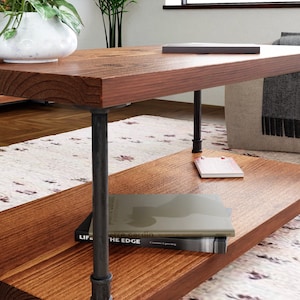 May include: A wooden coffee table with a lower shelf. The table has black metal pipe legs and a natural wood finish. The table is sitting on a rug with a floral pattern.