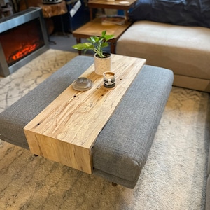 May include: A grey upholstered coffee table with a wooden top. The top is made of natural wood with knots and grain. A small potted plant and a cup of coffee are on the table.
