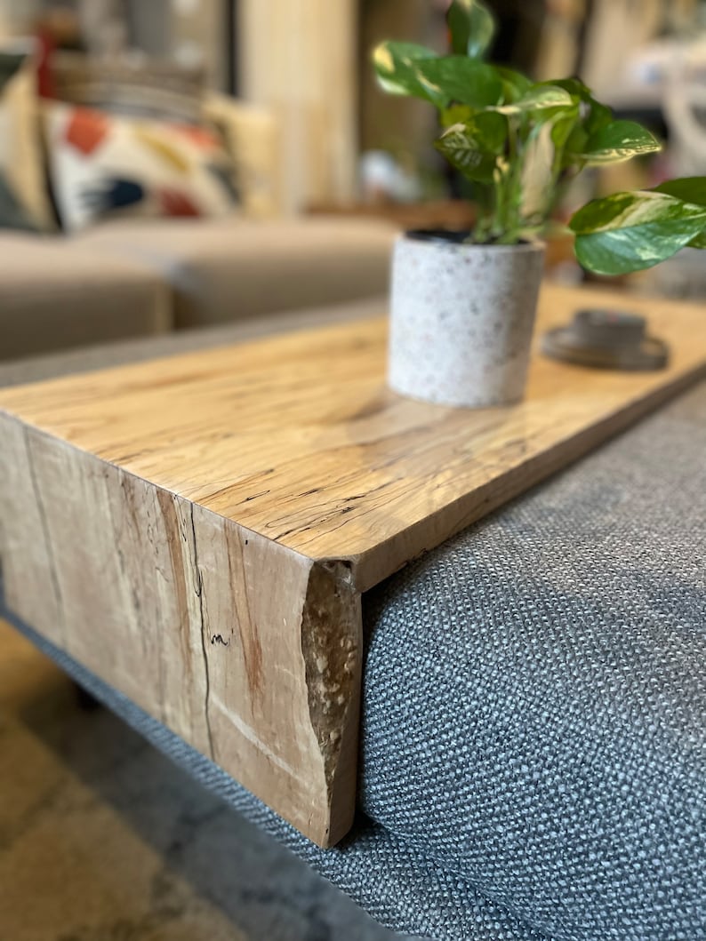 May include: A light wood side table with a live edge design. The table is placed on a grey sofa and has a potted plant on top.