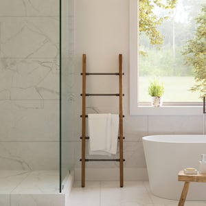 May include: A wooden towel ladder with dark metal rungs and a white towel in a modern bathroom. The bathroom features a white bathtub, a glass shower, and a window overlooking a green landscape. The walls are white with marble-like veining.