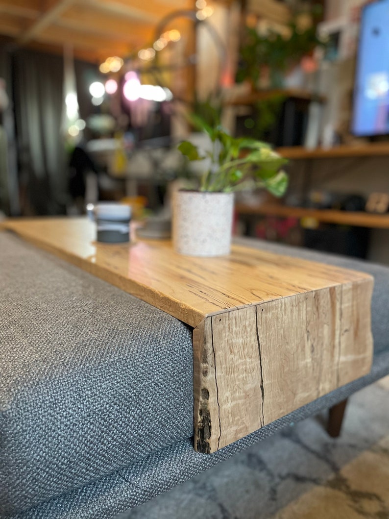 May include: A wooden tray with a natural wood finish sits on a grey sofa. The tray has a raised edge and is ideal for holding snacks or drinks.