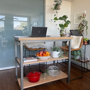 May include: A metal and wood kitchen island with two shelves. The top shelf has a laptop, a bowl of fruit, and a potted plant. The bottom shelf has a red pot, a glass cloche, and a cookbook titled "The Toronto Cooks".