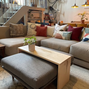 May include: A light gray ottoman with a wooden top coffee table. The ottoman and table are in a living room with a gray couch and colorful throw pillows.