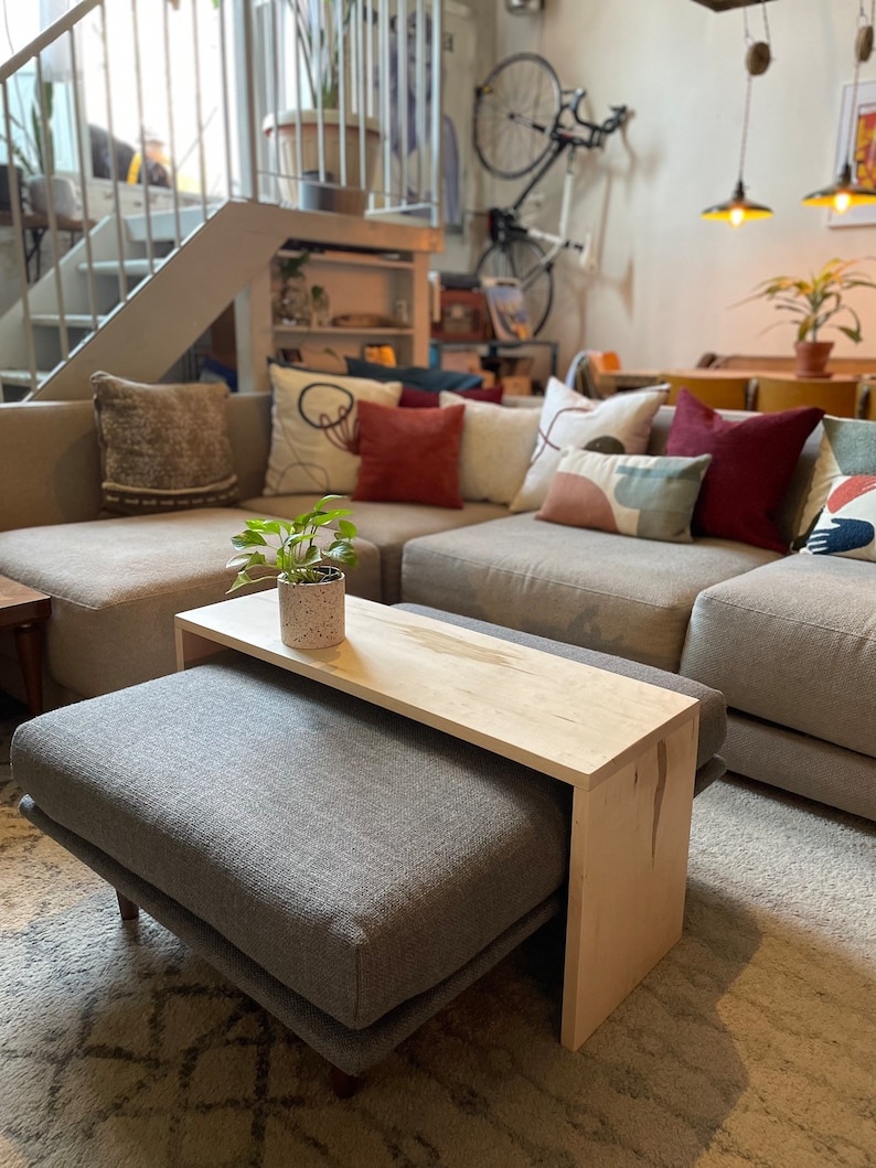 May include: A light gray ottoman with a wooden top coffee table. The ottoman and table are in a living room with a gray couch and colorful throw pillows.