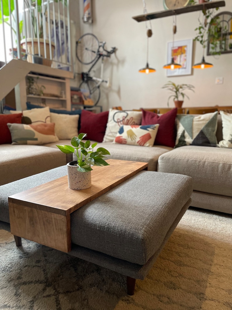 May include: A grey ottoman with a wooden tray attached to the top. The tray is made of light brown wood and has a small potted plant on it. The ottoman is sitting on a rug with a white and grey pattern.