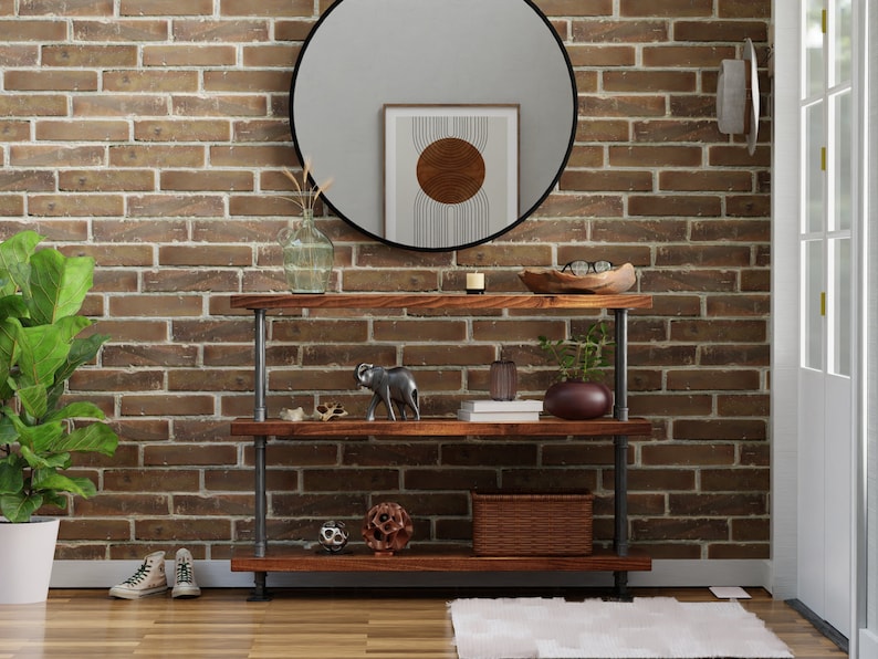 May include: A three-tiered industrial pipe shelf unit with wooden shelves. The shelf is against a brick wall and has various decorative items on it, including a small elephant figurine, a vase with dried flowers, a bowl with glasses, and a woven basket.