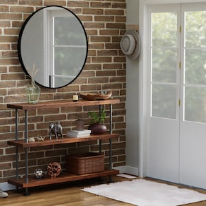 May include: A three-tiered wooden shelf unit with black metal pipe supports. The shelf is styled with decorative items, including a vase, a wooden bowl, books, and figurines. The shelf is placed in front of a brick wall with a round mirror hanging above it.