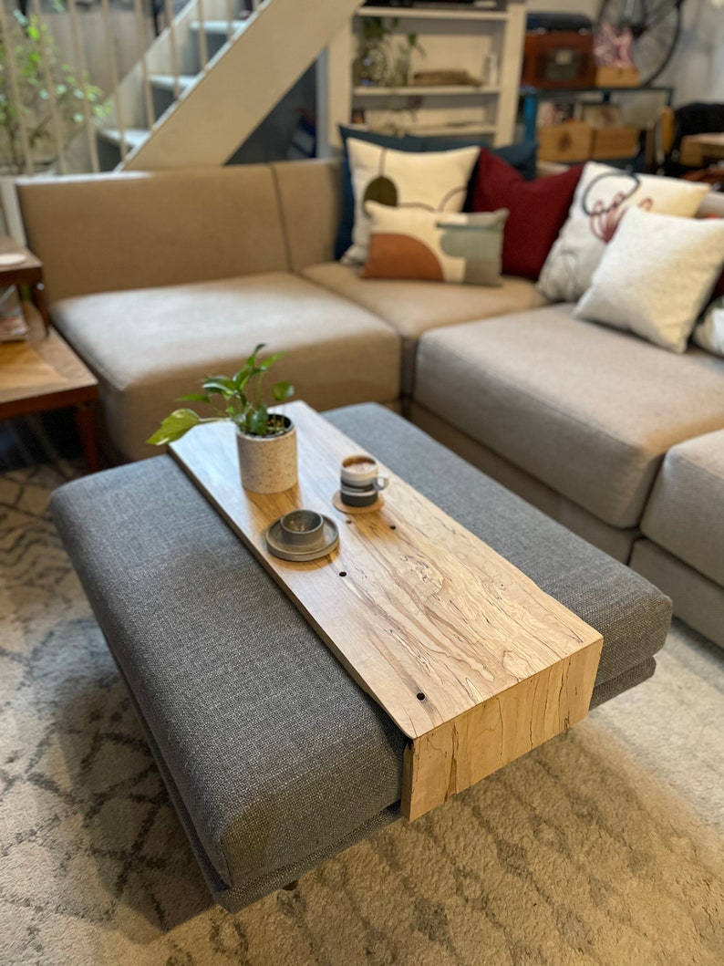 May include: A grey upholstered ottoman with a wooden top. The top is made of natural wood with a light brown finish. A small potted plant and two coffee cups are on the top.