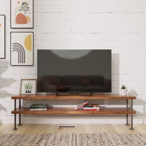 Puede incluir: Un soporte de TV de madera con dos estantes y patas de tubo de metal negro. El soporte tiene un televisor de pantalla plana, libros y una planta en maceta.