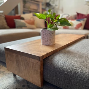May include: A wooden tray attached to a grey sofa with a potted plant on top. The tray is stained a dark brown colour and has a smooth, polished finish.