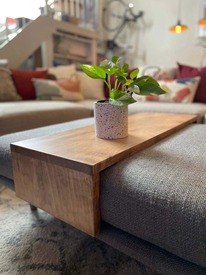 May include: A wooden tray attached to a grey sofa with a potted plant on top. The tray is stained a dark brown colour and has a smooth, polished finish.
