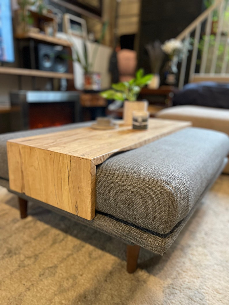 May include: A grey upholstered ottoman with a wooden top and a built-in shelf. The ottoman has a grey fabric cover and wooden legs.