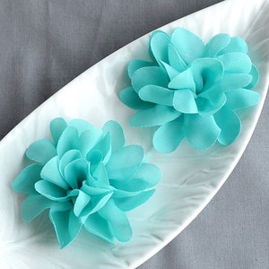 May include: Two turquoise fabric flower embellishments with layered petals, displayed on a white, leaf-shaped dish. The flowers have a soft, textured appearance and are set against a gray background.
