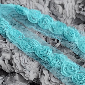 May include: Two rows of turquoise blue fabric roses sewn onto a sheer, light blue mesh fabric. The fabric is textured and has a delicate, floral pattern.