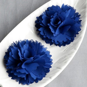 May include: Two vibrant blue fabric flower embellishments rest on a white, leaf-shaped dish. The flowers have a layered petal design, creating a full, textured appearance. The background is a soft gray, providing a neutral backdrop.