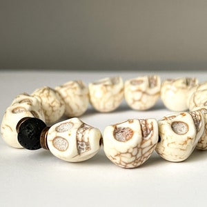 May include: A white skull bead bracelet with a black lava bead. The bracelet is made of white beads with a brown marbled pattern.