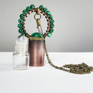 May include: A copper and brass necklace with a green malachite stone pendant and a glass roller bottle. The necklace has a chain and a circular design with green malachite beads.
