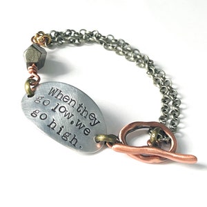 May include: A silver bracelet with a charm that reads "When they go low, we go high." The bracelet has a copper toggle clasp and a pyrite bead.