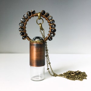 May include: A copper and brass necklace with a glass vial pendant. The vial is topped with a black and white stone and a copper cap. The necklace is made with a chain and a circle of black and white stones.