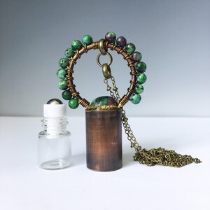 May include: A copper and brass pendant necklace with a green and pink gemstone, surrounded by a circle of green beads. The necklace is attached to a small, clear glass bottle with a roller ball.