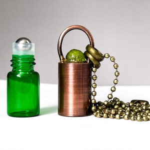 May include: A green glass roller bottle with a silver roller ball next to a copper-colored metal roller bottle with a green glass bead and a gold chain.